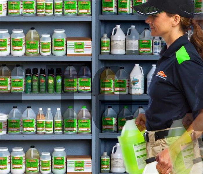 A woman in SERVPRO gear holding a gallon of green product while standing in front of a wall of product